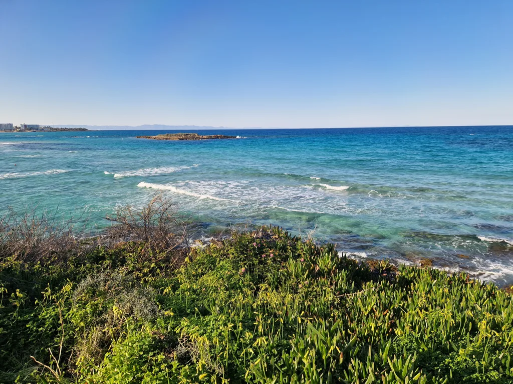 Shallow, clear turquoise water over pale sand at Fig Tree Bay, Protaras, on the east coast of Cyprus, with headlands and mountains on the horizon.